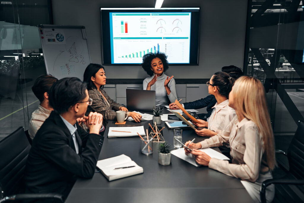 Young confident successful business people discuss and analyze work, statistics, plans, startup in a meeting room with a display screen behind them. Image: HBS]/stock.adobe.com