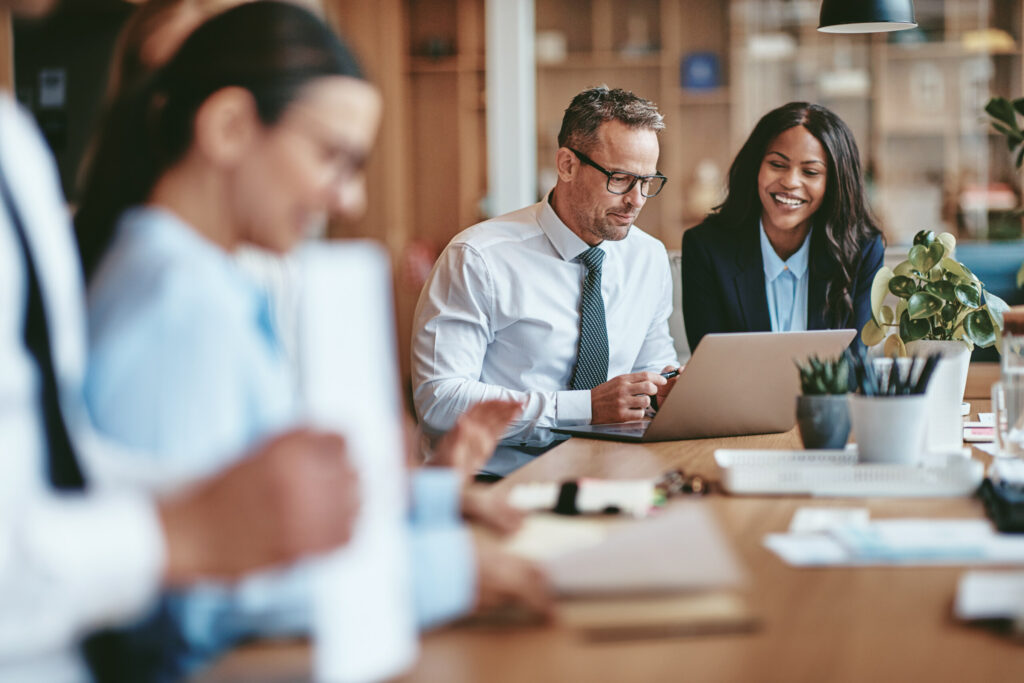 Two diverse businesspeople smiling while working on a laptop together at the end of a boardroom table in an office.
