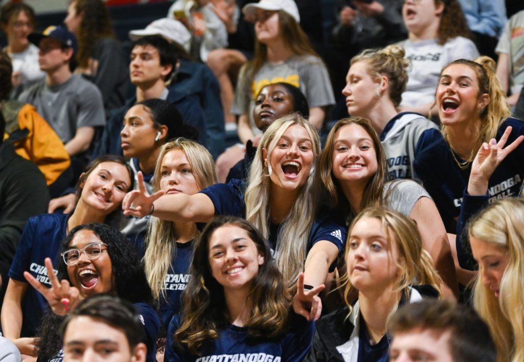 Students in navy shirts cheering at basketball game.