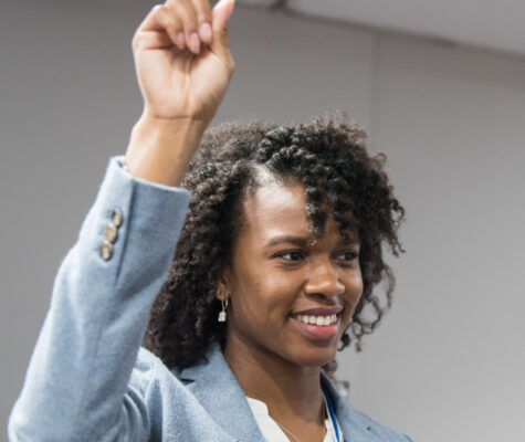 Young woman in a light blue suit raising her hand.