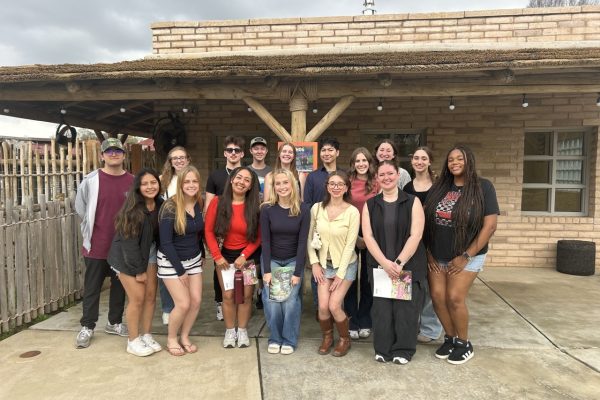 A group of 16 students pose for a group photo in front of a rustic building with a thatched roof. Students are arranged in two rows and smile at the camera on a cloudy day.