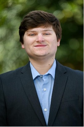 A young man is smiling, standing in front of trees