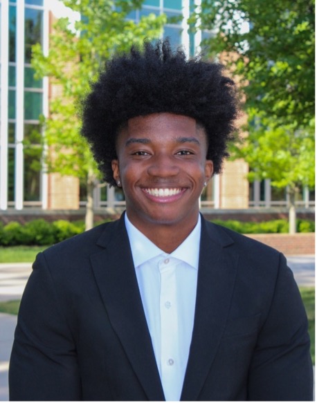 A young man is smiling, standing in front of a brick and glass building