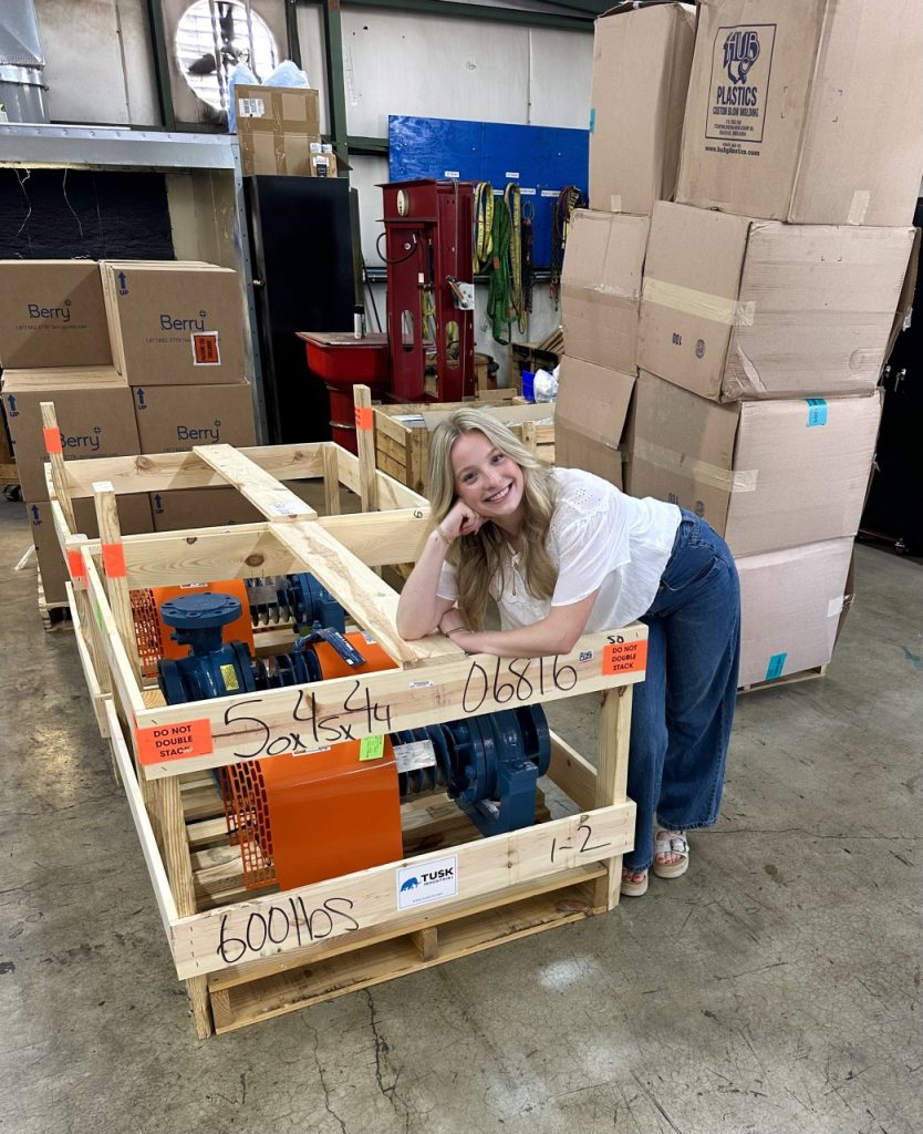 Kristin Lyle playfully leans over a pallet of machinery. 
