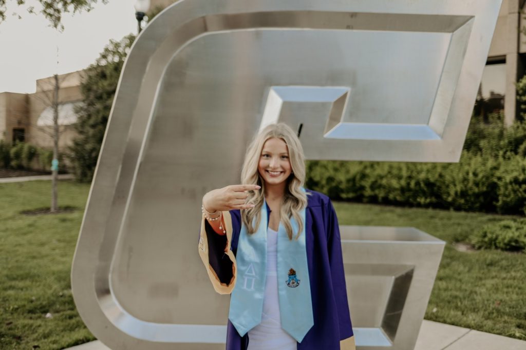 Kristin Lyle poses in her graduation gown on campus at UTC showing a Power C with her hand. 
