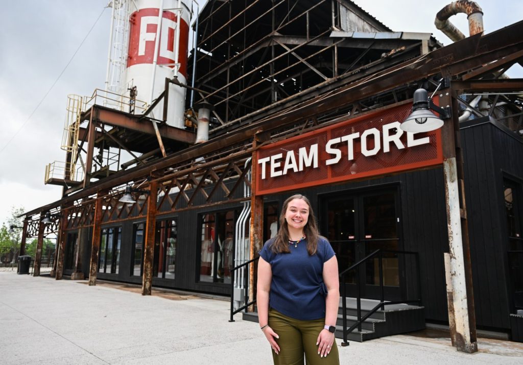 Woman wearing blue shirt stands in front of a sign that says Team Store