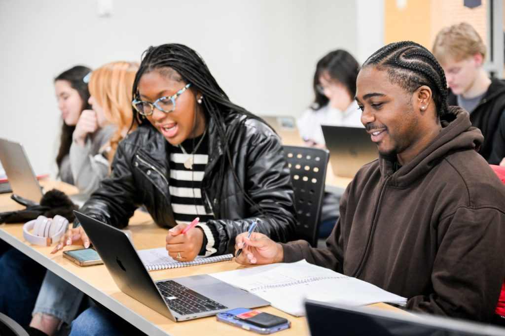 Woman and man sit at desk for discussion. Woman is writing. Man is looking at a computer screen.