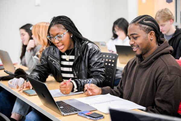 Woman and man sit at desk for discussion. Woman is writing. Man is looking at a computer screen.