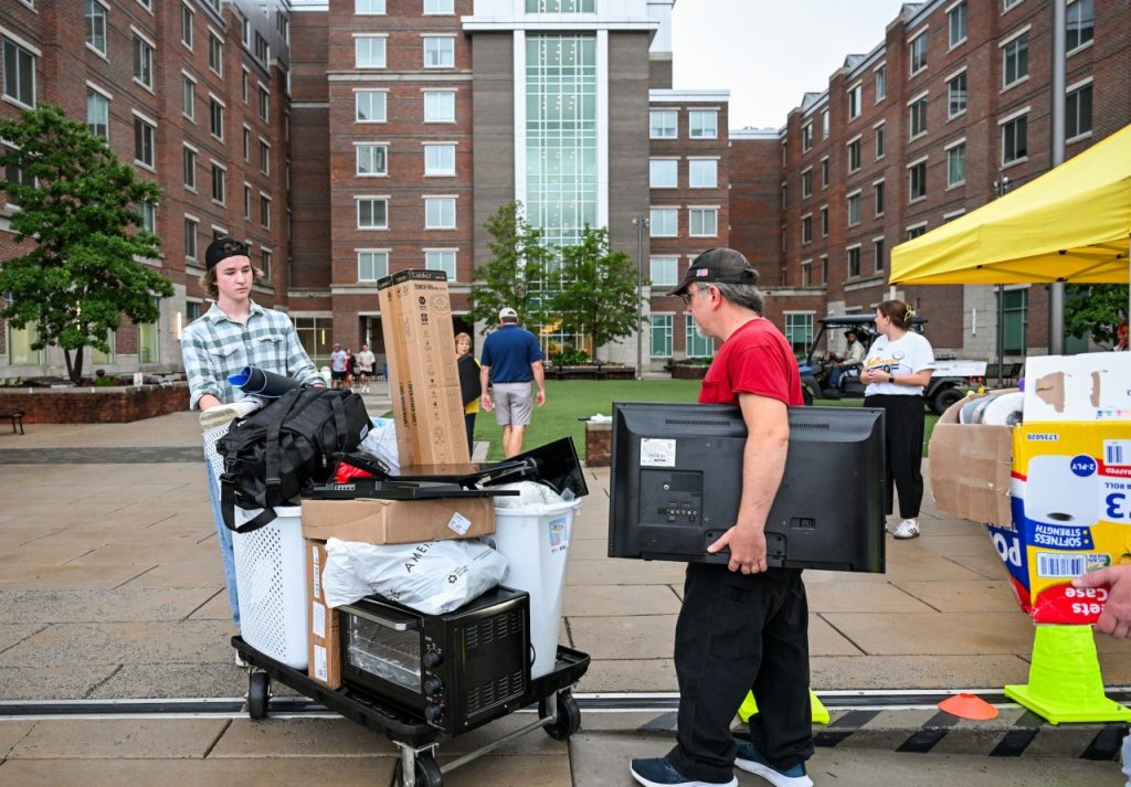 Student pushes cart with boxes, while man follows carrying TV