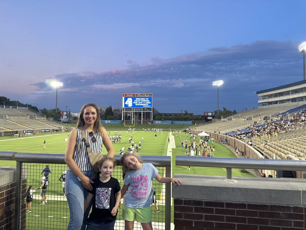 A mom and two daughters smile in front of a football field