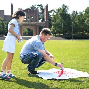 Trevor Elliott kneels on Chamberlain Field as he helps a Girls Preparatory School camper prepare a model rocket for launch. The two stand beside the rocket on its launch pad with Founders Hall visible in the background.