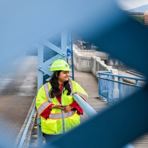 UTC graduate student Nishat Tasnim Nisha, wearing a hard hat and reflective safety vest, leans on the railing of Chattanooga's Market Street Bridge. She looks toward the river as the bridge's blue steel framework creates a geometric frame around her.