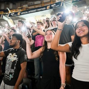 Students smile while they hold cellphone flashlights in the stands at Convocation