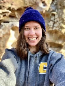 Amy is holding the camera to take a smiling selfie. She is wearing a blue beanie and a grey UTC hoodie. There are rock formations behind her in the background.