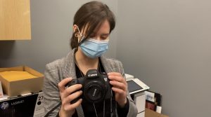 Amy is wearing a medical mask and is holding a camera while taking a mirror selfie.