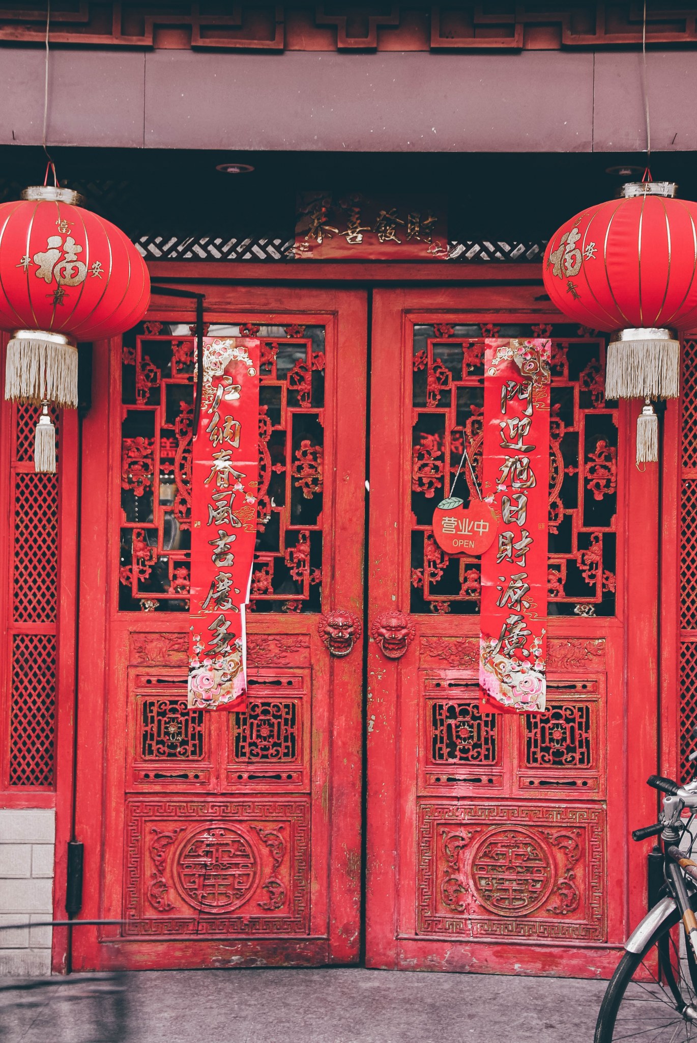 Large red wooden doors with red paper tapestries and lanterns hanging in front. One on each side.