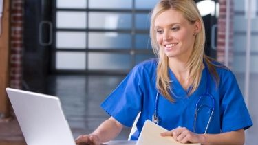 blonde woman wearing scrubs and looking at a computer while holding files