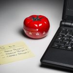 a tomato timer sitting between a black laptop and a to do list