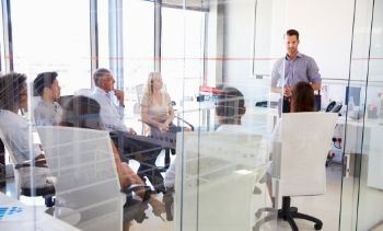 man leader standing up talking to his team members who are sitting down in a conference room