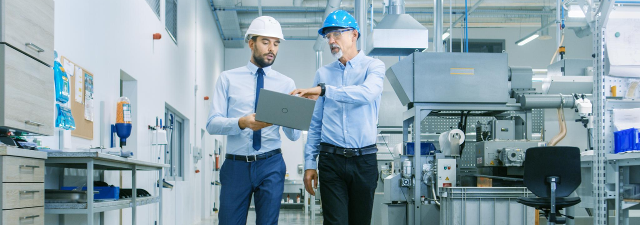 two male supply chain workers looking at a laptop while walking through a warehouse