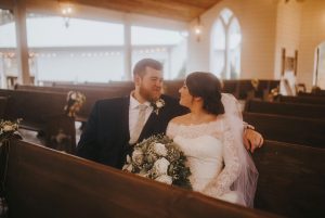 Sydney and Casey in a pew of the wedding chapel, gazing adoringly at each other in wedding gown and tux