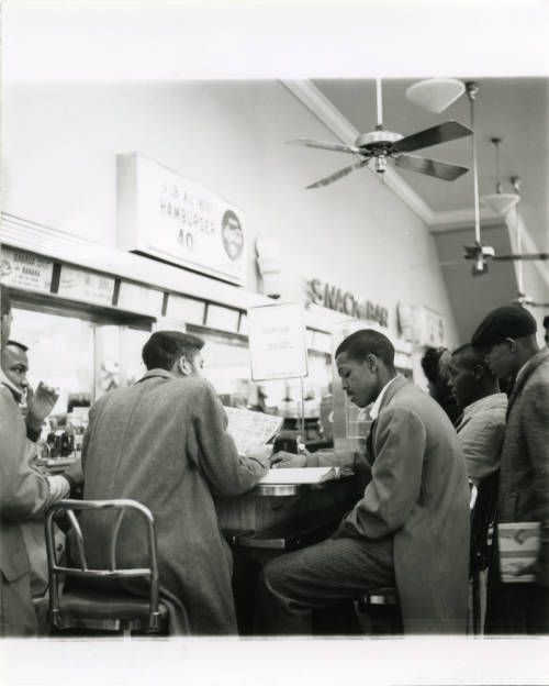 	
Black-and-white photograph of Black students from Howard High School protesting at a lunch counter sit-in in downtown Chattanooga, Tennessee. A sign on the counter reads, "FOUNTAIN CLOSED."