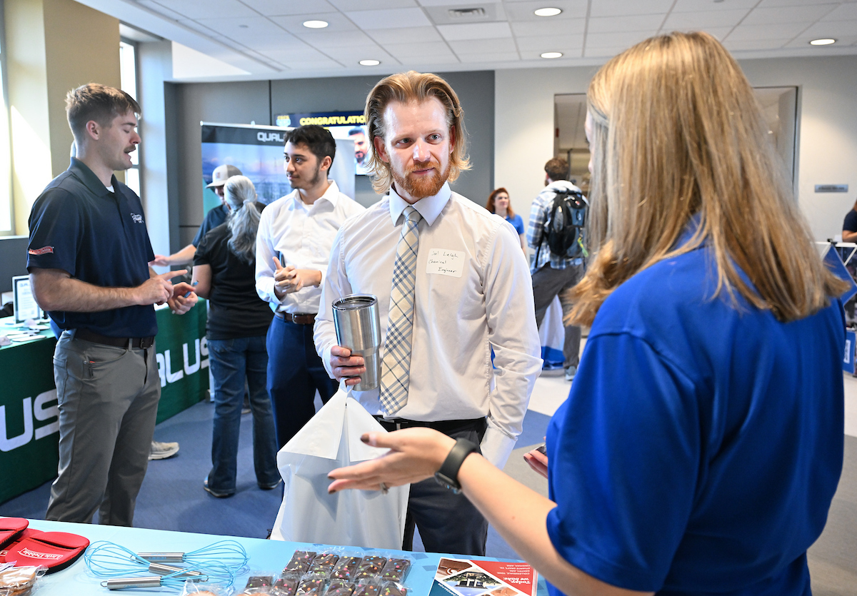Joel Leigh is holding a cup and a bag, listening to a recruiter. 