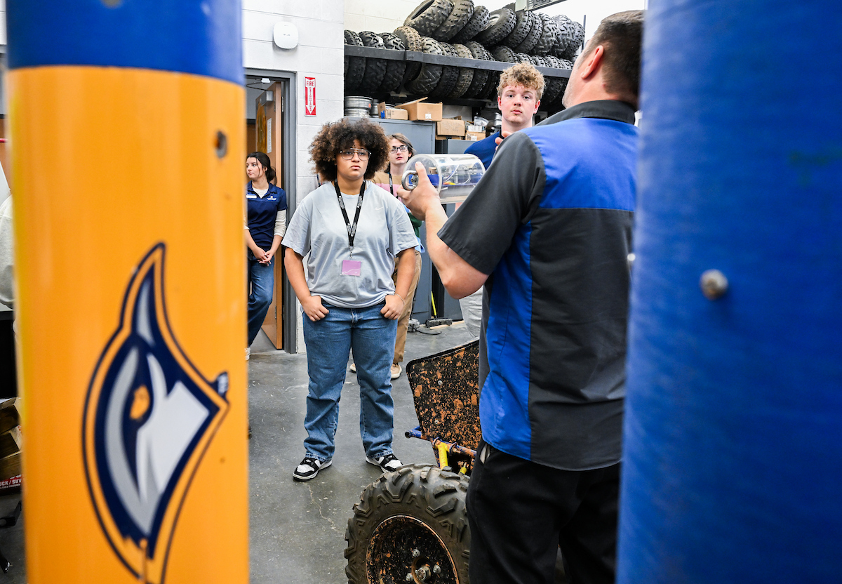 A group of students listens as an instructor gives a demonstration in a workshop filled with equipment and stacked tires.