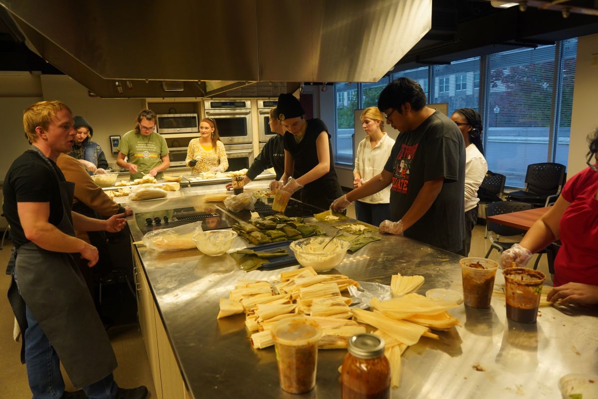 A group of students and community members work together in a commercial kitchen to make tamales, standing around a large stainless steel counter covered with masa, corn husks, fillings, and jars of ingredients during a hands-on workshop.