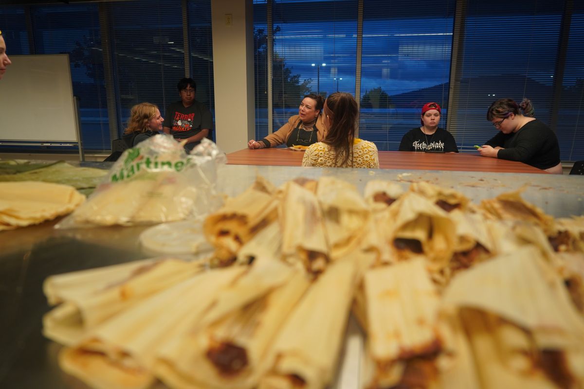 A group of students sit around a table talking in a classroom at dusk, with a large tray of freshly made tamales in the foreground out of focus.