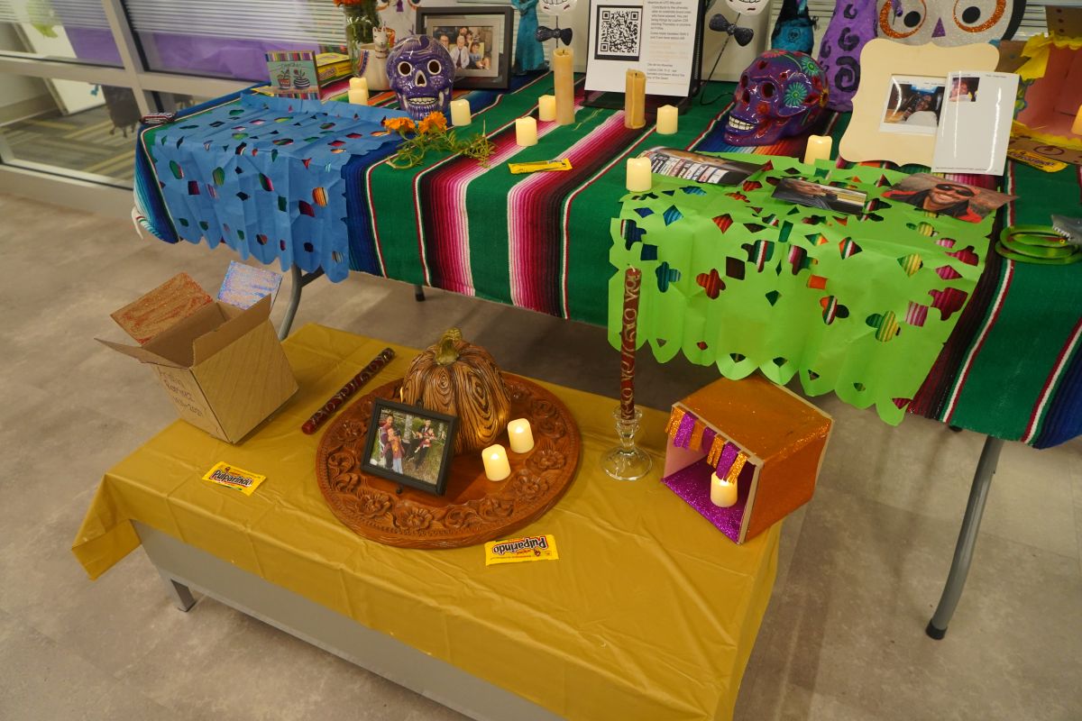 A Día de los Muertos ofrenda display features colorful papel picado, decorated sugar skulls, candles, family photos, flowers and small personal items arranged on vibrant cloth-covered tables.