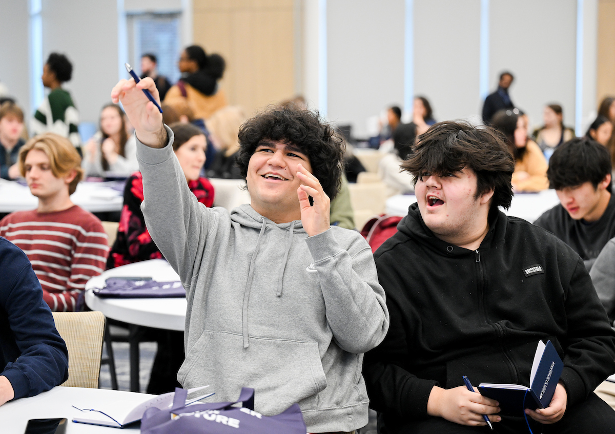 A student in a grey hoodie smiles and points at the screen while a student in a black hoodie looks where he’s pointing. 