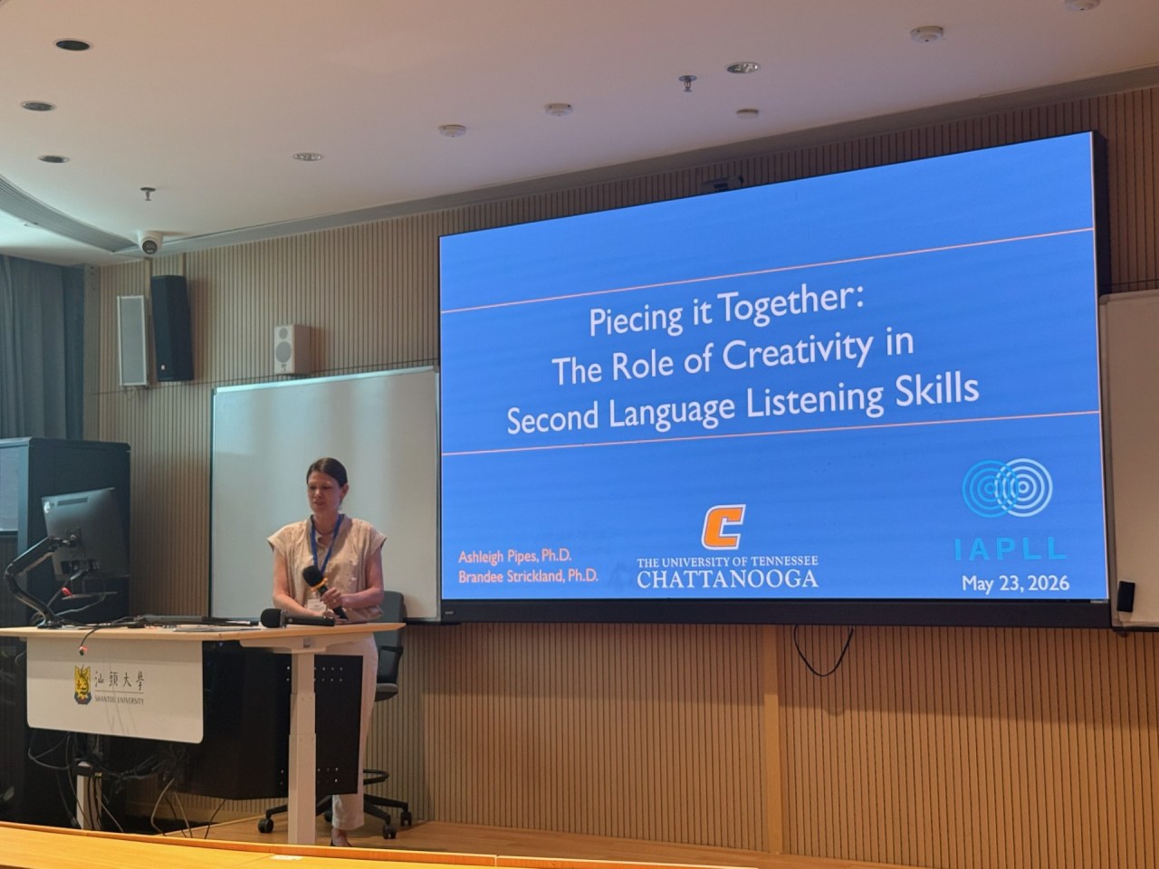 Dr. Ashleigh Pipes stands at a lectern presenting during the International Association for the Psychology of Language Learning conference at Shantou University in China. A large presentation screen displays the title, "Piecing it Together: The Role of Creativity in Second Language Listening Skills," with UTC and IAPLL logos.